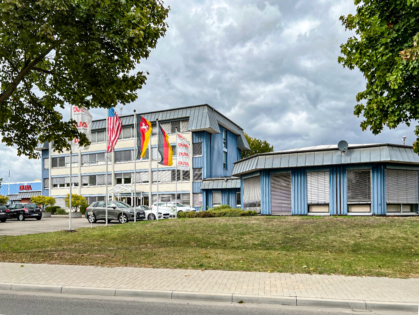 Blaues Bürogebäude mit Fahnen auf Parkplatz, Rasenfläche und Bürgersteig im Vordergrund
